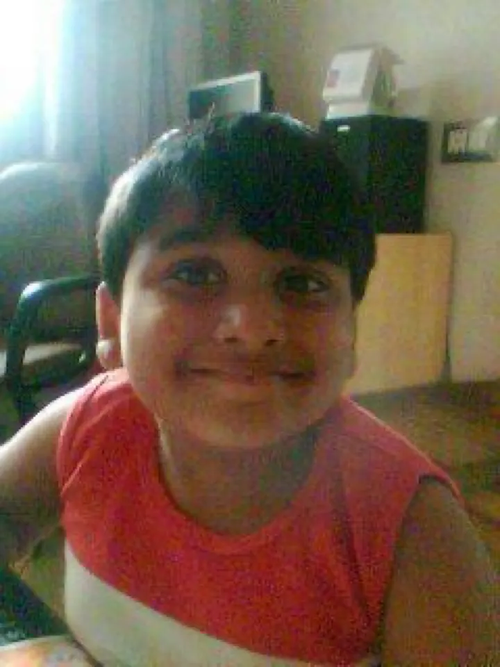 Me as a boy in a red and white T-shirt, grinning at the camera, a desk and a curtained window behind me.