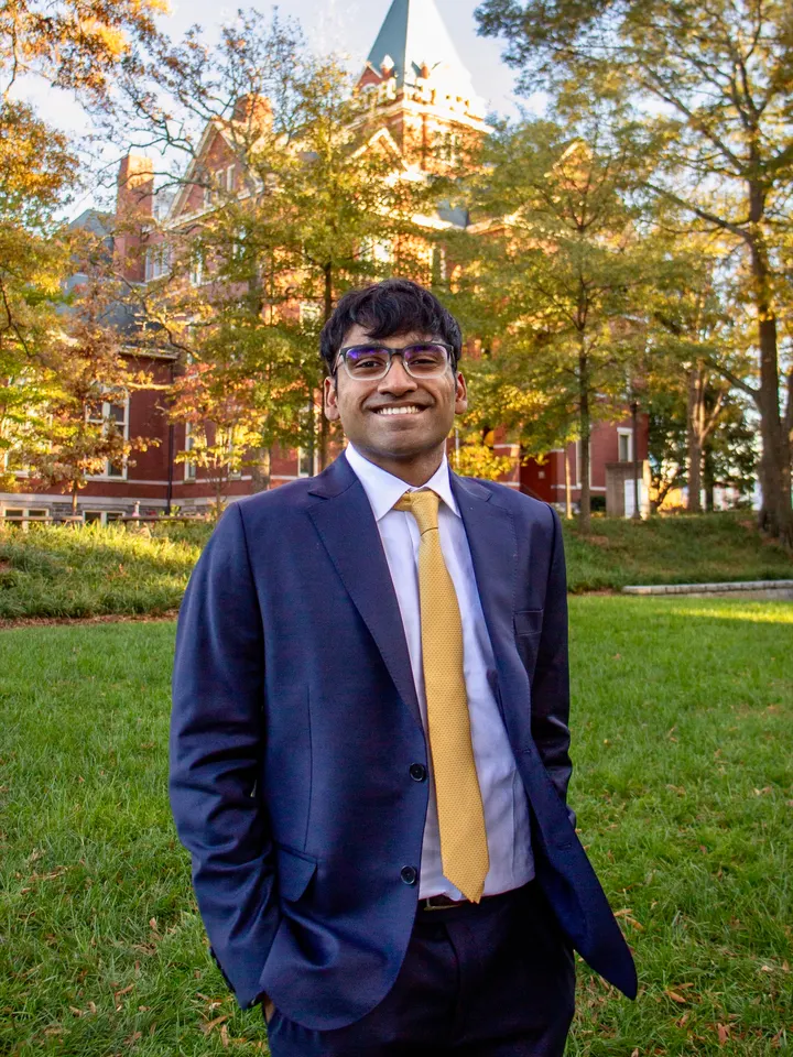 Me in a navy suit and gold tie, hands in my pockets, on a lawn at Georgia Tech, with red brick, a spire and autumn trees behind me.