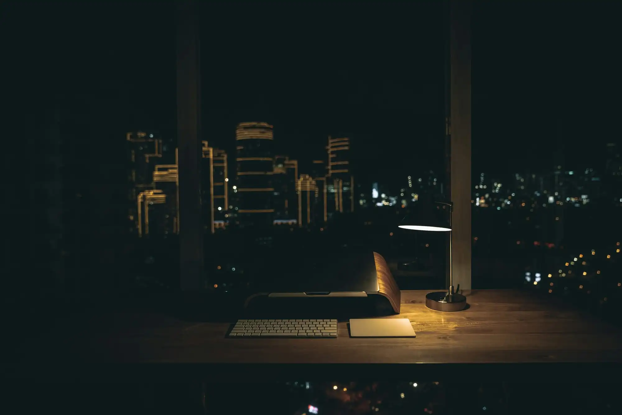 A desk at night: a lamp lights a keyboard, and city lights show through the window behind it.