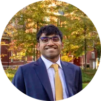 Saahil Sanganeriya, smiling, in a suit, in front of autumn trees and a brick building.
