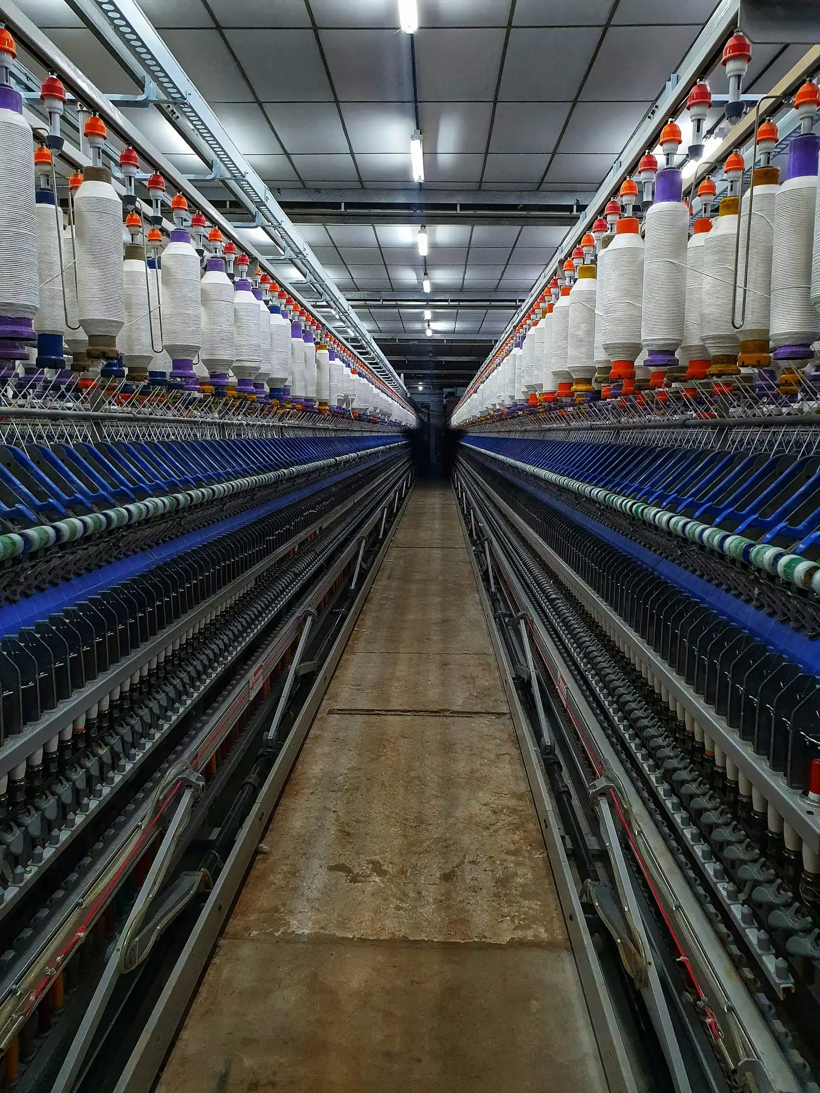 A long aisle between ring-spinning frames in a mill, with rows of white bobbins on both sides.